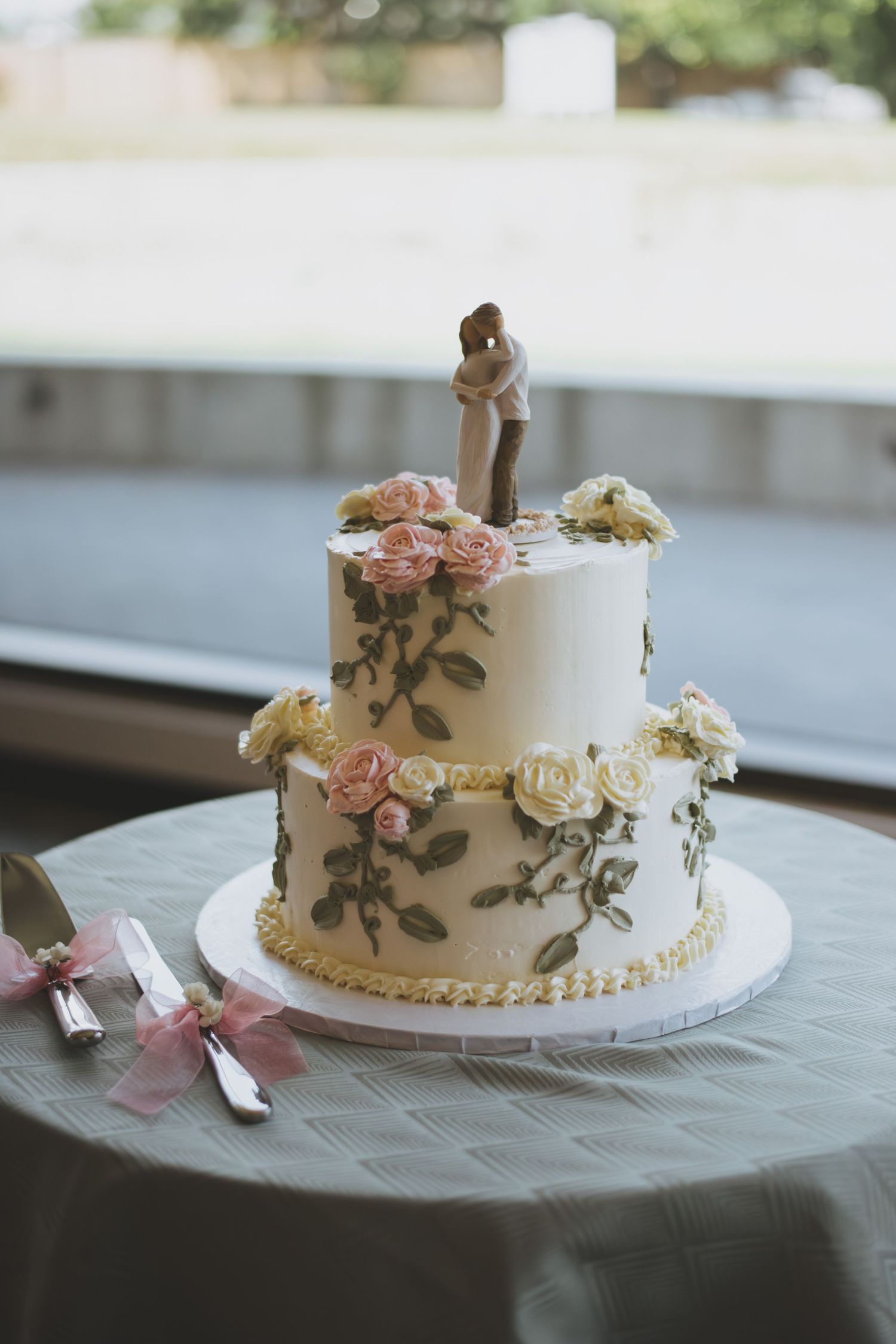 Two-tier white wedding cake decorated with delicate pink and cream roses and gray floral designs on a table.
