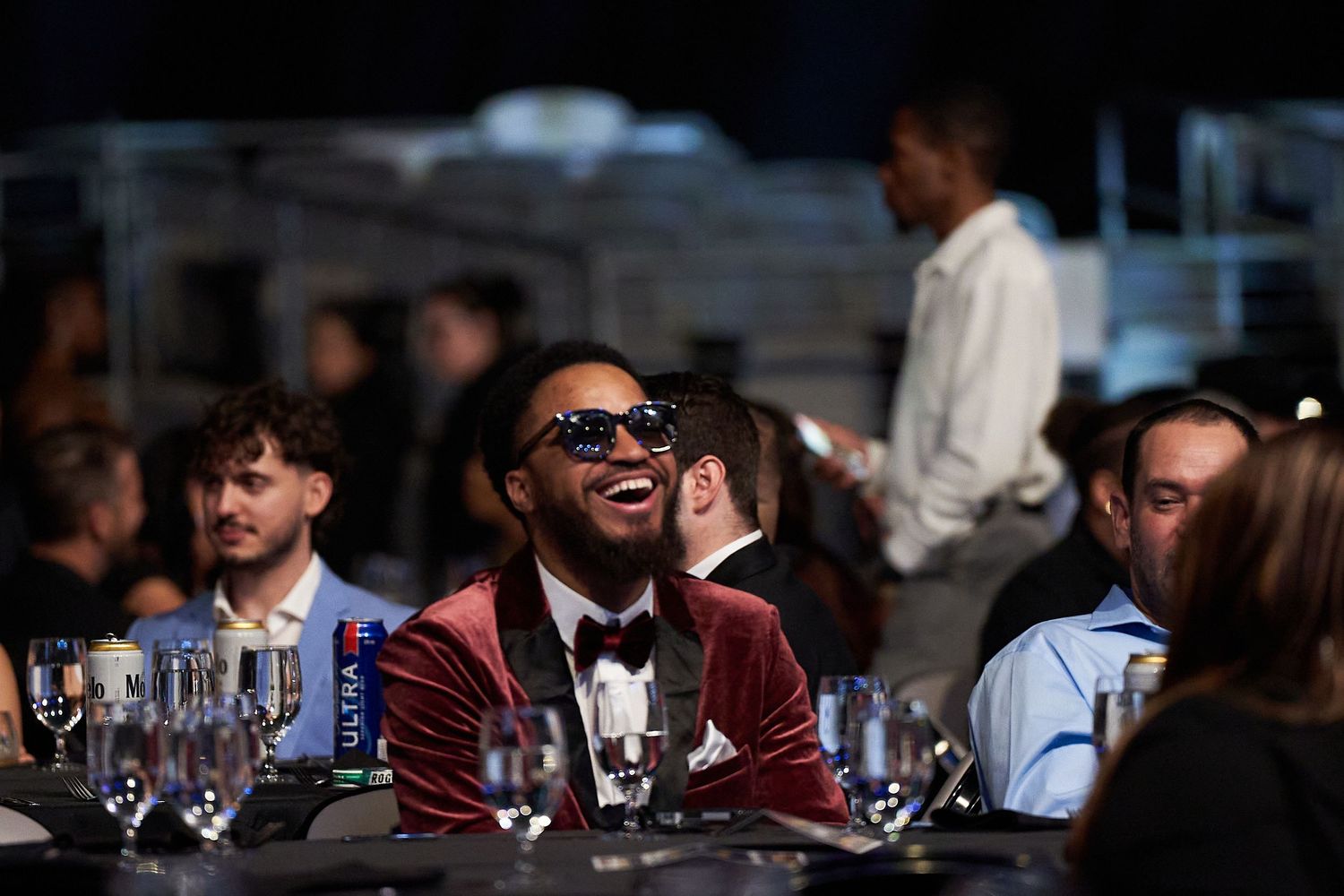 Person in burgundy velvet jacket and sunglasses laughing while seated at formal event with table settings.