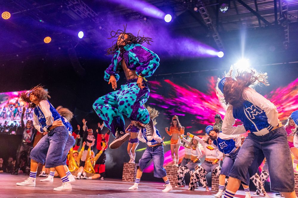 Dynamic dance performance with performers in vibrant costumes leaping and moving under purple stage lighting.