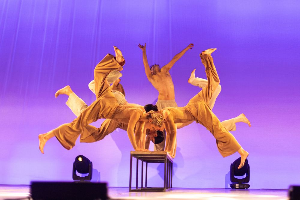 Modern dancers in fluid motion perform against purple-lit stage backdrop.