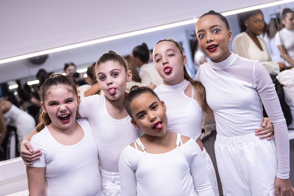 Young ballet dance students in white leotards making silly faces while posing together at a dance studio.