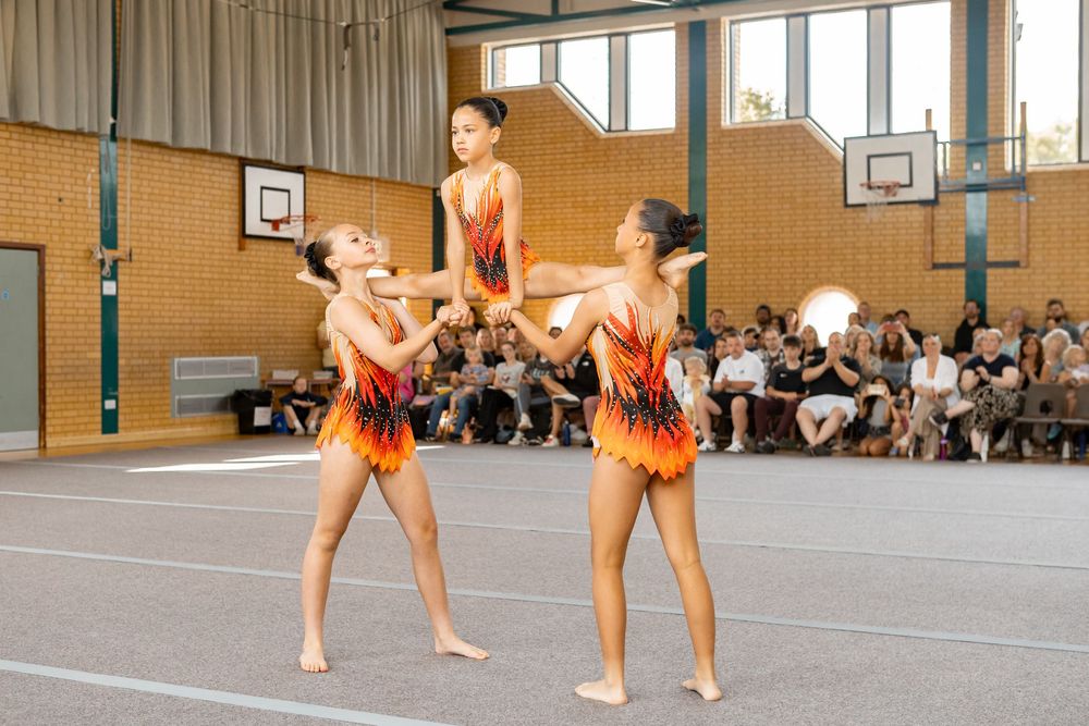 Three young gymnasts in orange leotards perform an acrobatic routine at an indoor competition with spectators watching.