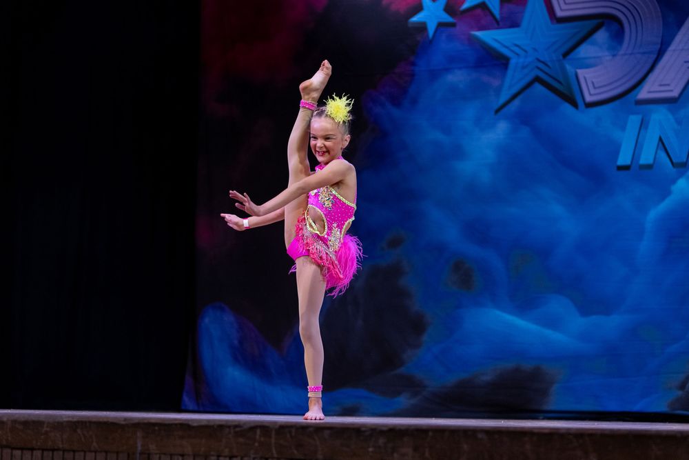 Young dancer in bright pink costume performs energetic dance move on stage with blue starry backdrop.