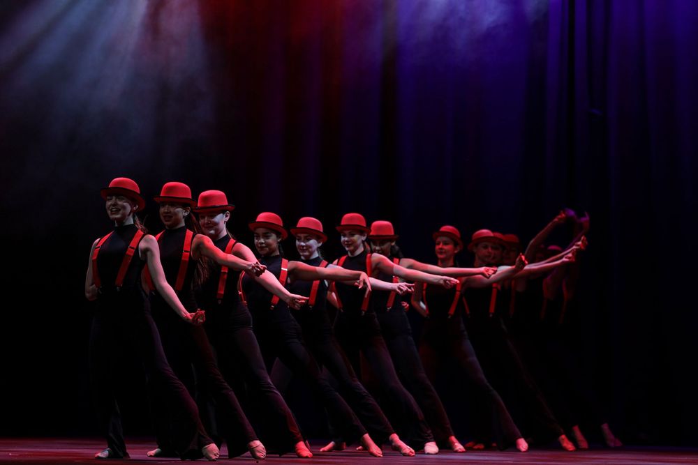 Dancers in red caps perform synchronized lean movement on dark stage with dramatic lighting and shadows.
