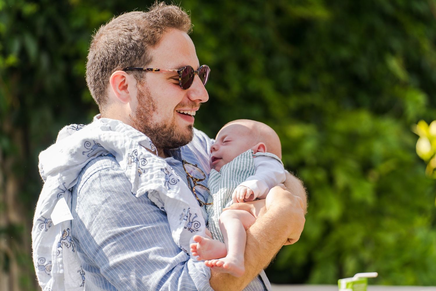 A person wearing sunglasses and a striped button-down shirt holds a small infant while standing outdoors on a sunny day.