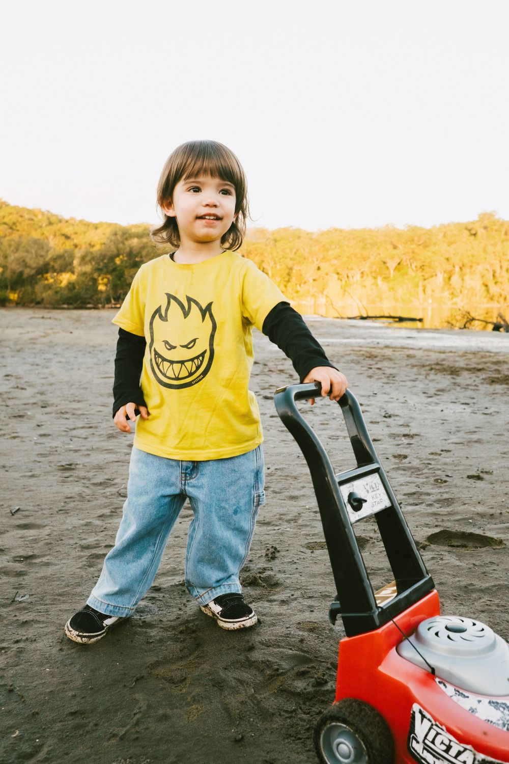 Child in yellow t-shirt playing with red toy lawnmower on beach.