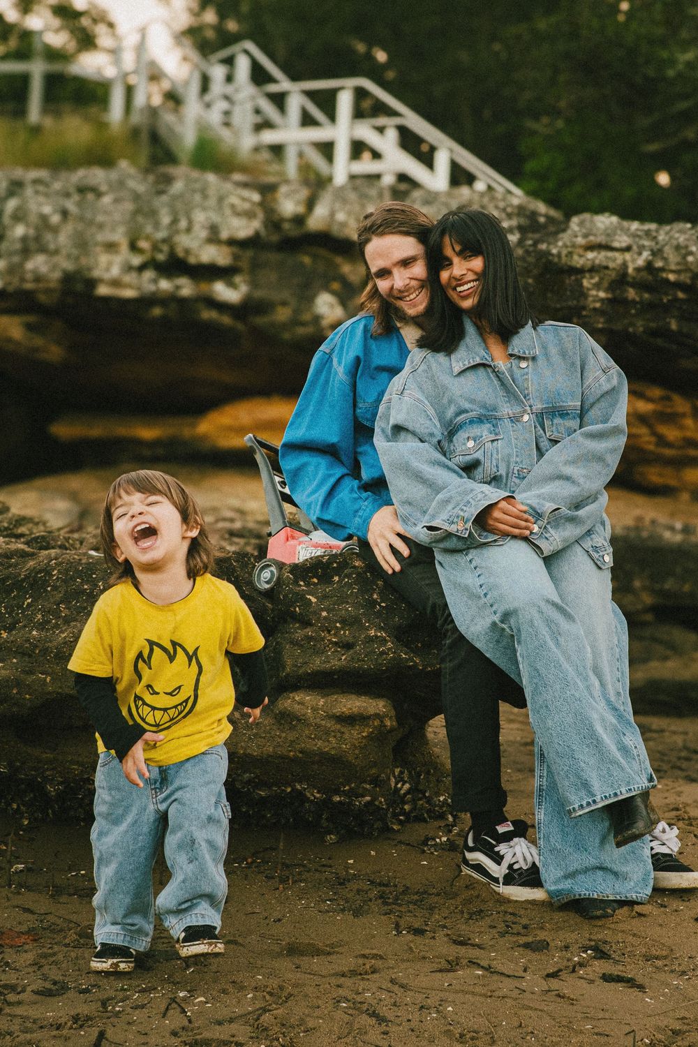 Family sitting on rocky beach with child in yellow smiley face t-shirt laughing.