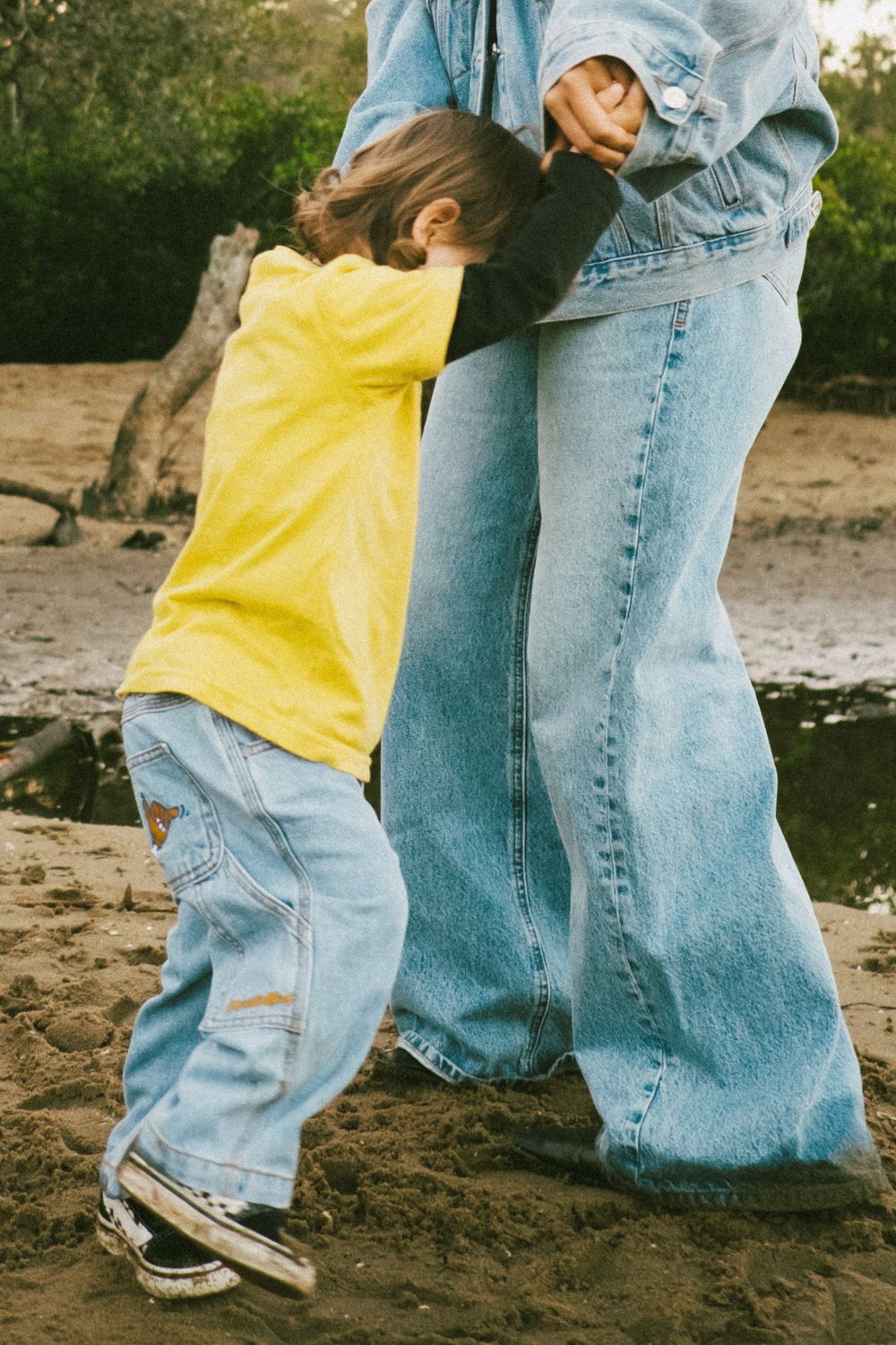 Young child in yellow shirt hugging someone wearing denim jeans on a beach.
