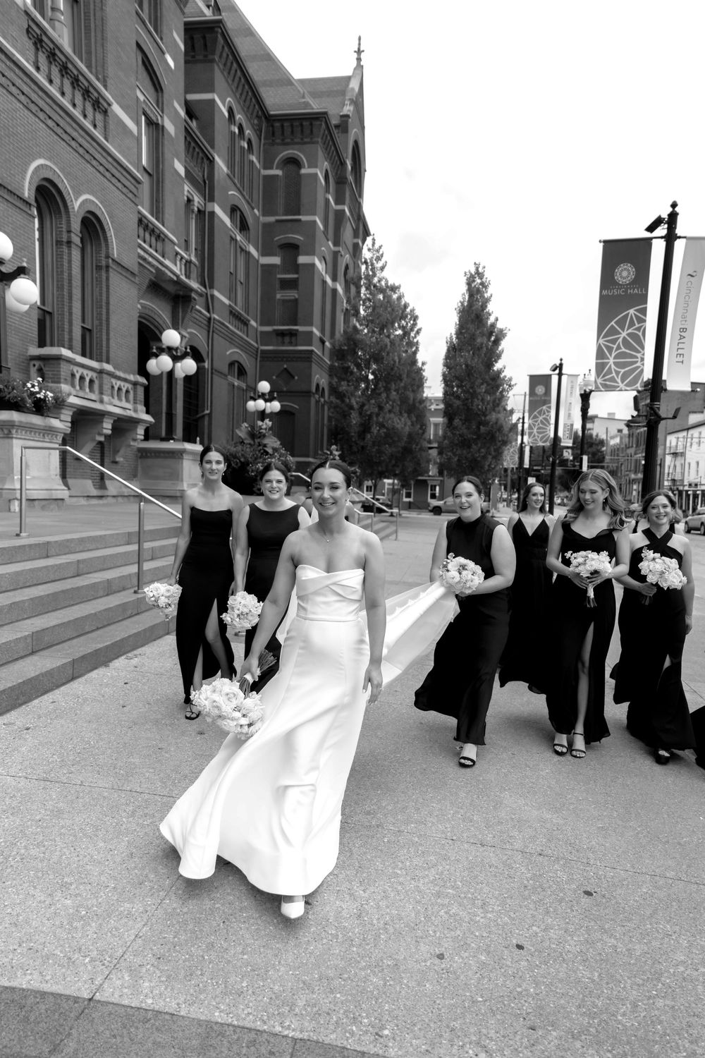 Wedding party in black attire walks down a city sidewalk alongside historic brick buildings.