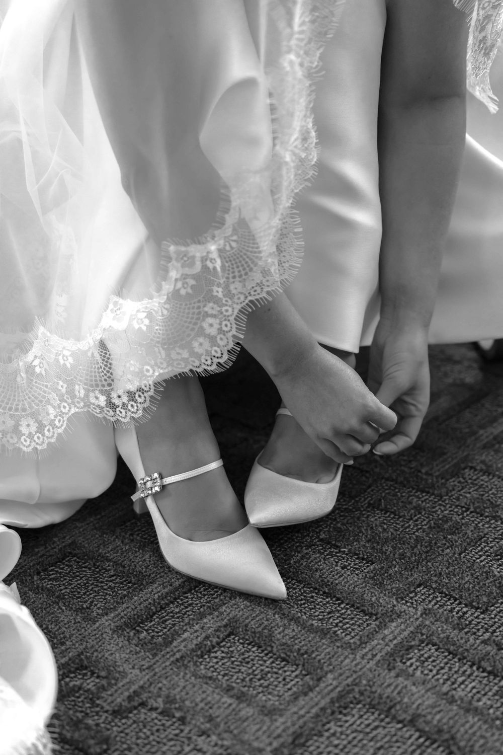 Close-up black and white photo of bride adjusting white wedding shoe with lace dress hem visible.