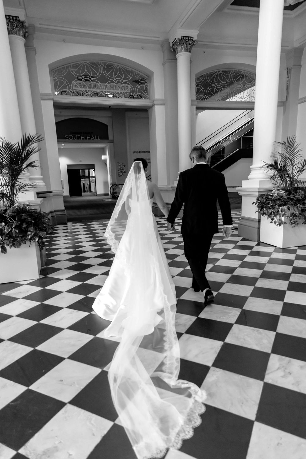A bride and groom walk across an elegant black and white checkered floor with ornate columns and flowing veil trailing behind.