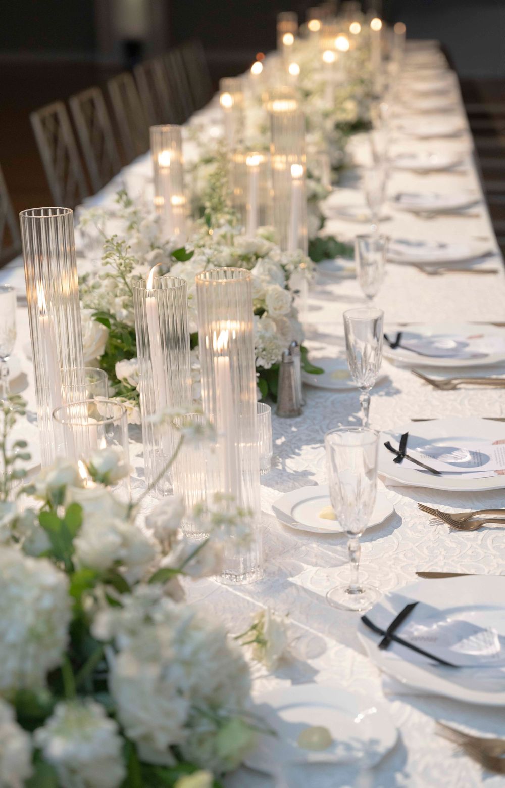 Elegant white wedding table setting with glass candleholders and white floral centerpieces along a long banquet table.