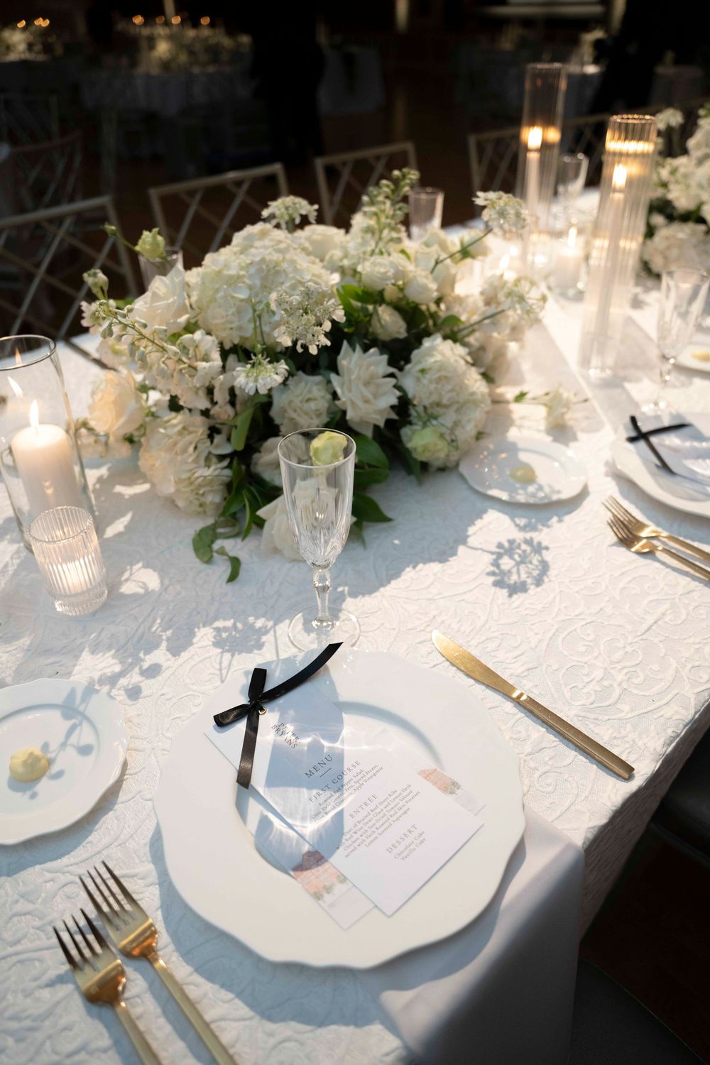 Elegant white wedding reception table setting with floral centerpiece, candles and place settings on white tablecloth.