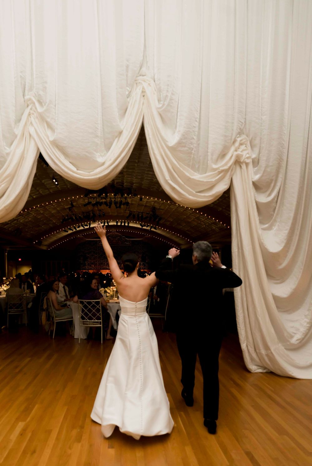 A couple dances at a wedding reception under elegant white draped fabric decorations on a hardwood dance floor.