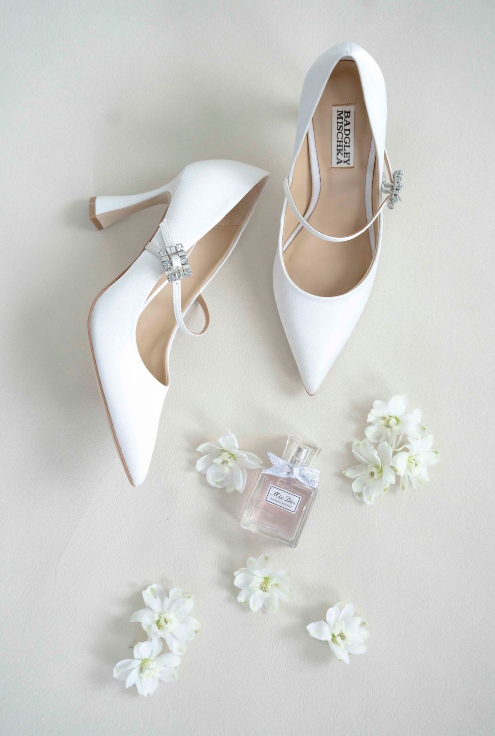 Elegant white bridal shoes arranged with delicate white flowers and perfume bottle on light neutral background.