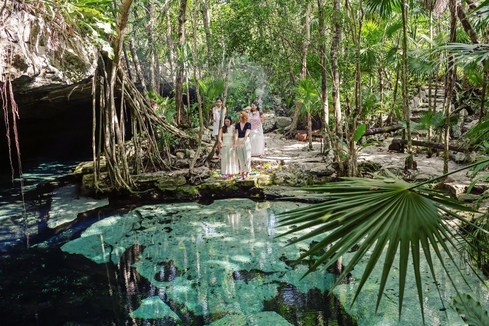 People stand beside a crystal clear cenote surrounded by lush jungle vegetation and hanging vines in Mexico.