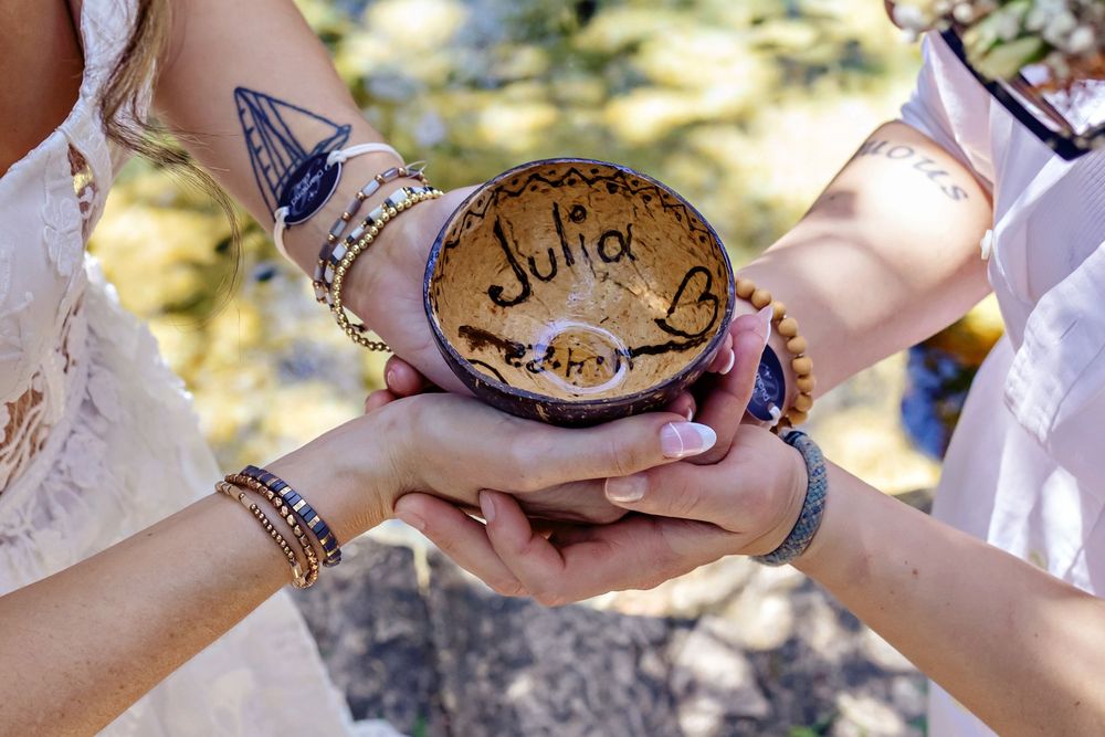Multiple hands coming together holding a rustic wooden bowl with decorative carvings at an outdoor ceremony.