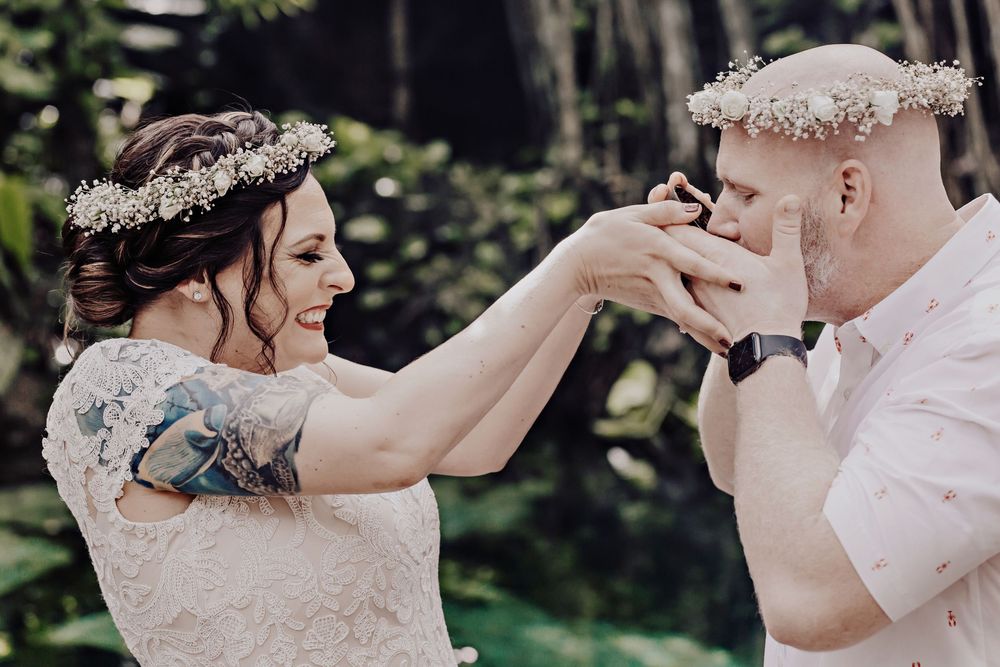 Wedding couple wearing floral crowns share a tender moment outdoors as the groom kisses the bride's hand.