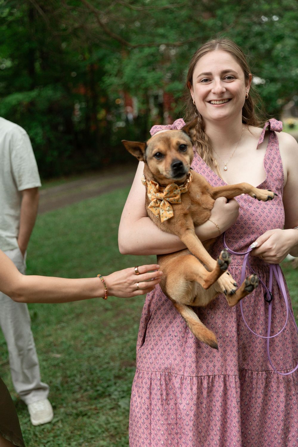 Bridesmaid in pink floral dresses holding small brown dog at an outdoor wedding.