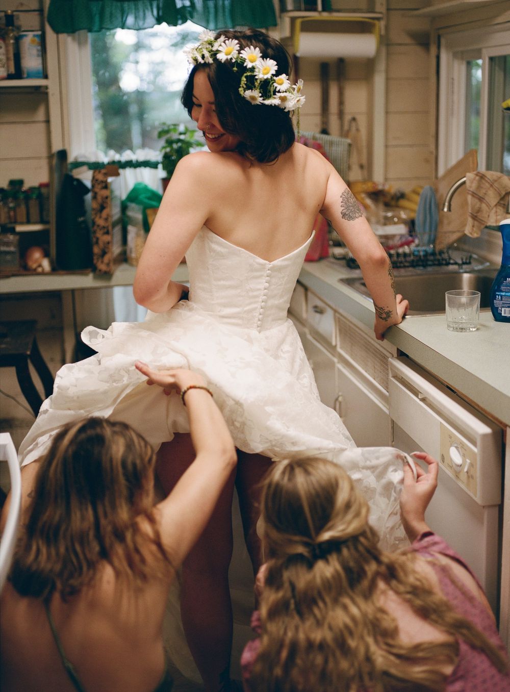 A bride in a white wedding dress with a flower crown leans against a kitchen counter while others help with her dress.