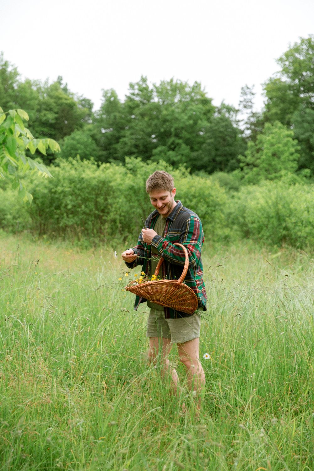 Person in plaid shirt walking through tall grass field with woven basket.