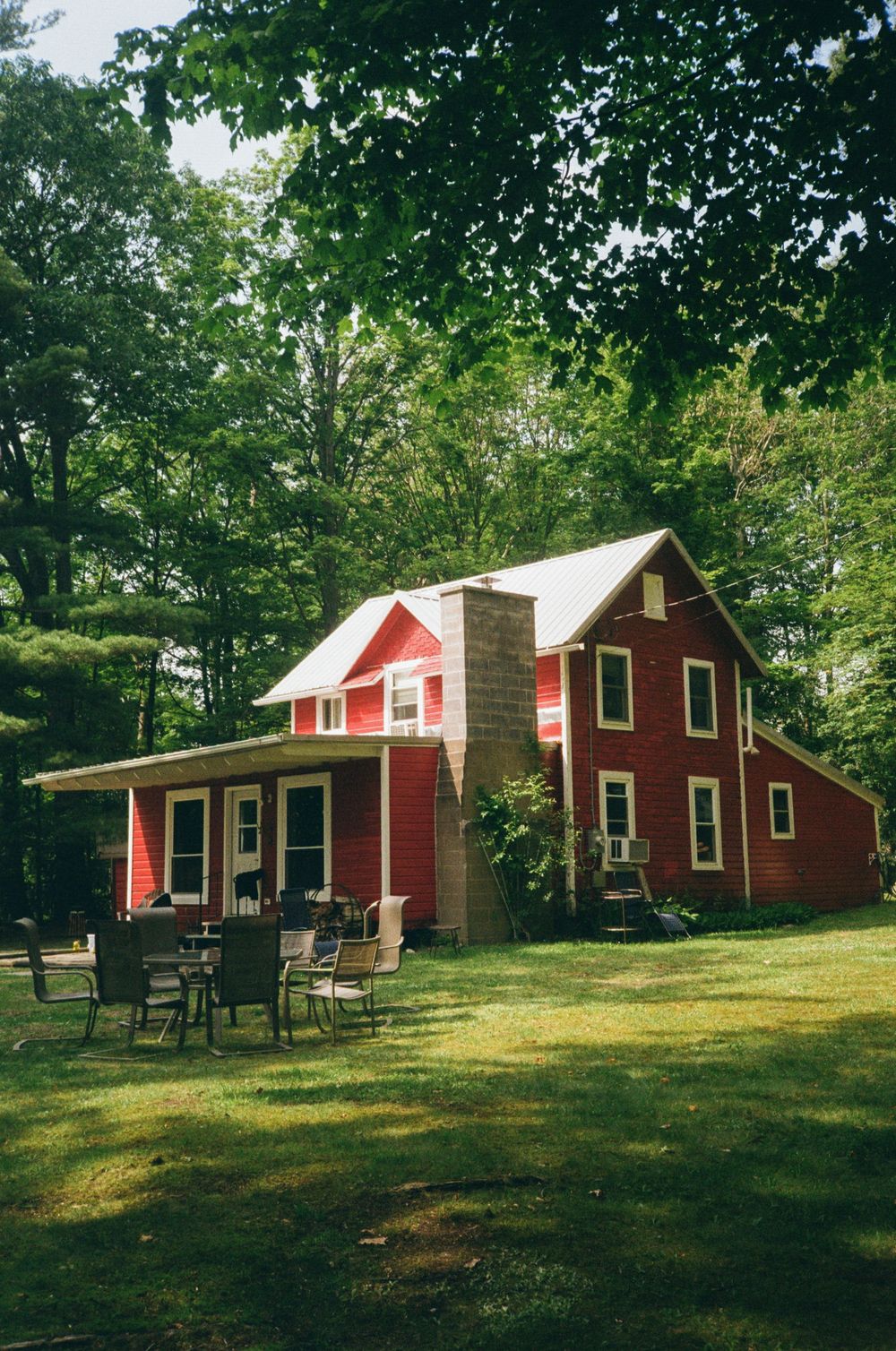 A charming red farmhouse with white trim nestled in the woods with outdoor seating on the lawn.