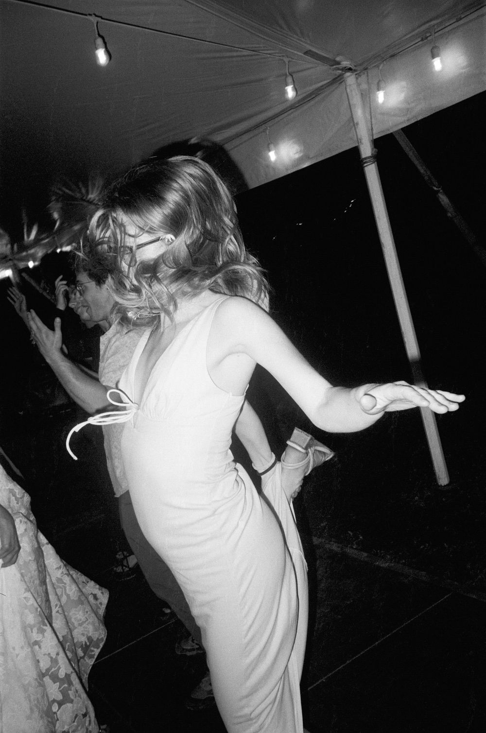 A guest in a white dress dances energetically under a tent at an evening wedding reception.