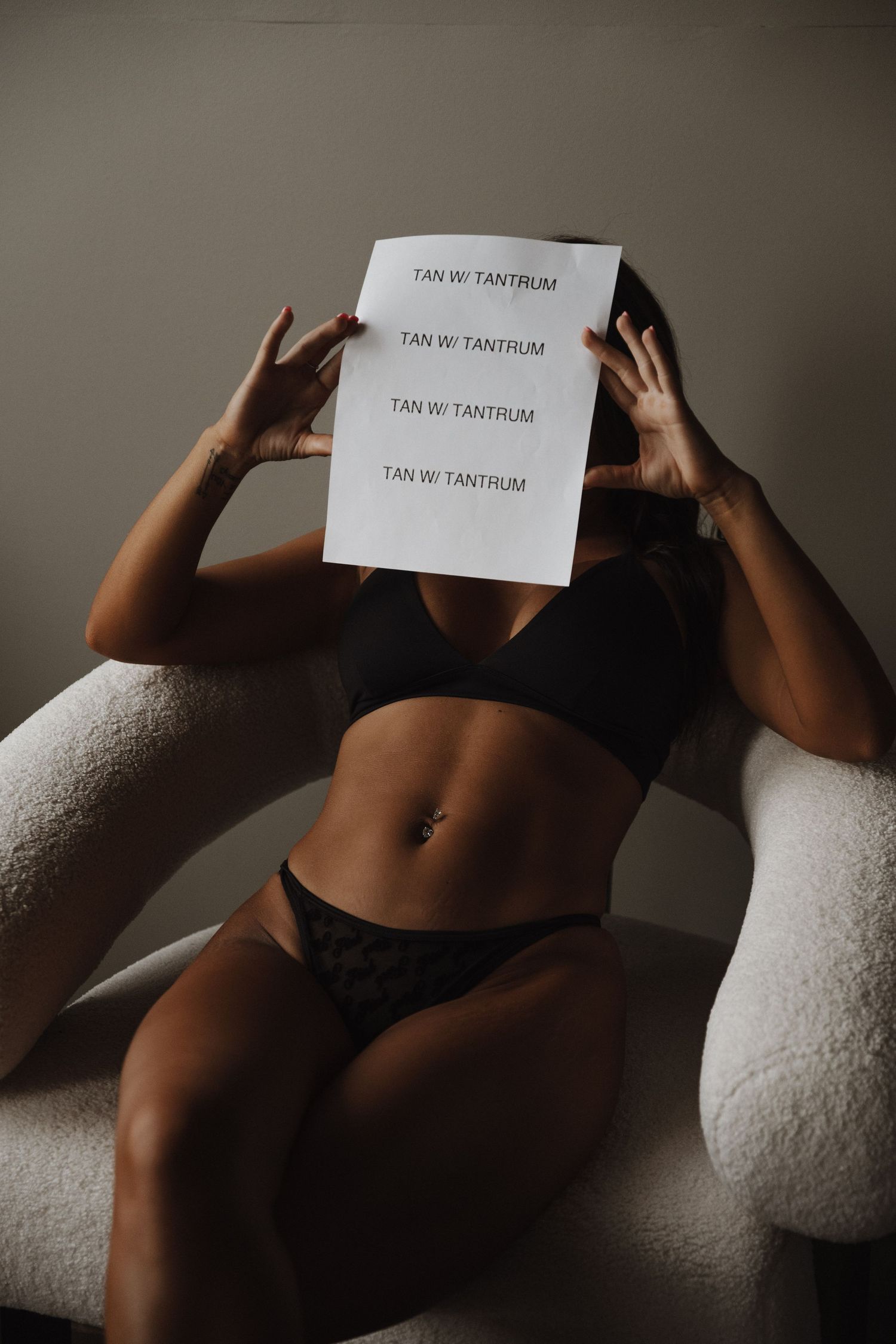 Person in black lingerie lounging on white chair holding paper with repeated text 'I am NE Tantrum' against moody lighting.