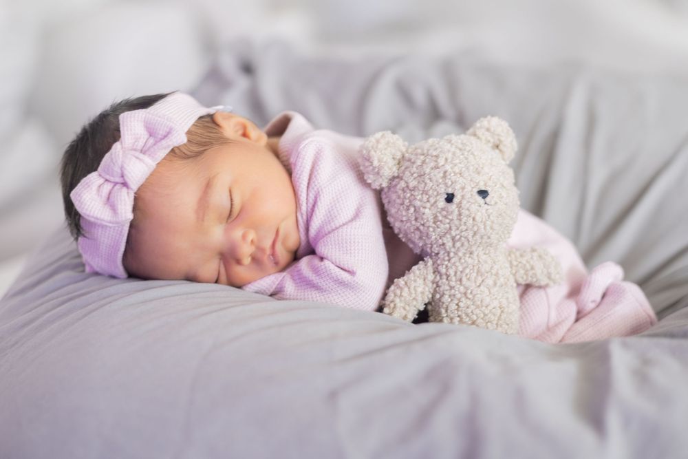 A sleeping baby dressed in pink clothing lies on soft gray bedding with a plush toy companion.