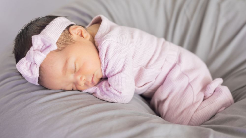 A peaceful sleeping newborn baby wearing soft pink clothing and headband rests on gray bedding.