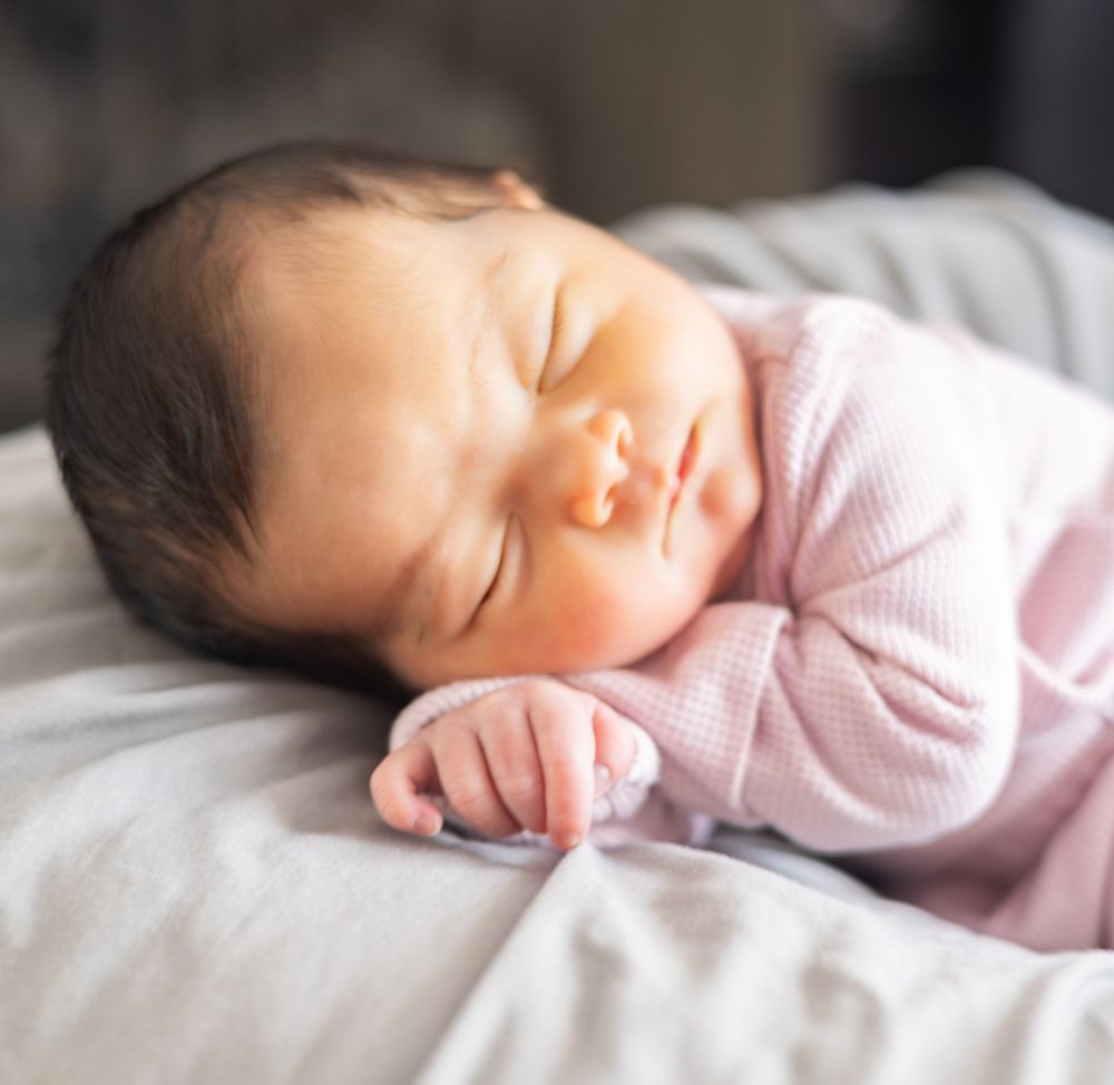 A sleeping newborn baby wearing soft pink clothing rests peacefully on white bedding.