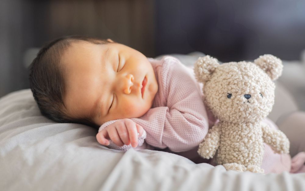 Peaceful sleeping infant in pink clothing rests beside a plush teddy bear on white bedding.