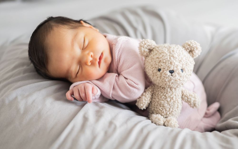 A peaceful baby in pink sleepwear rests on white bedding next to a plush teddy bear companion.