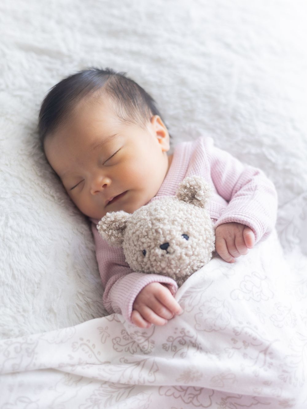 A sleeping newborn in pink clothing cuddles with a soft teddy bear on white bedding.