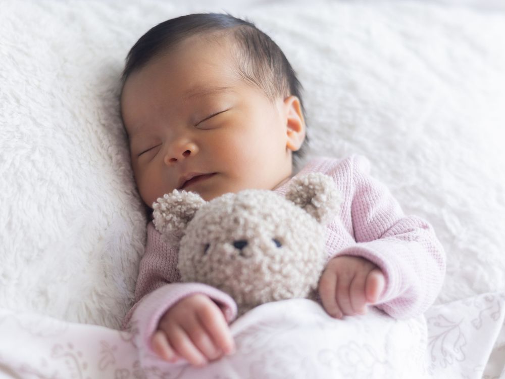 Baby wearing a pink outfit sleeps peacefully on white blankets while holding a small plush toy.