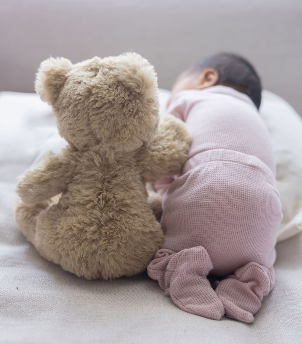 Baby wearing pink clothing lies next to a soft beige teddy bear on white bedding.