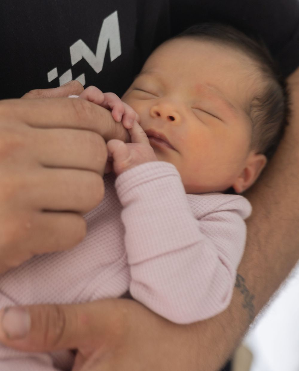 A sleeping newborn baby swaddled in a pink blanket is being gently held by caring hands.