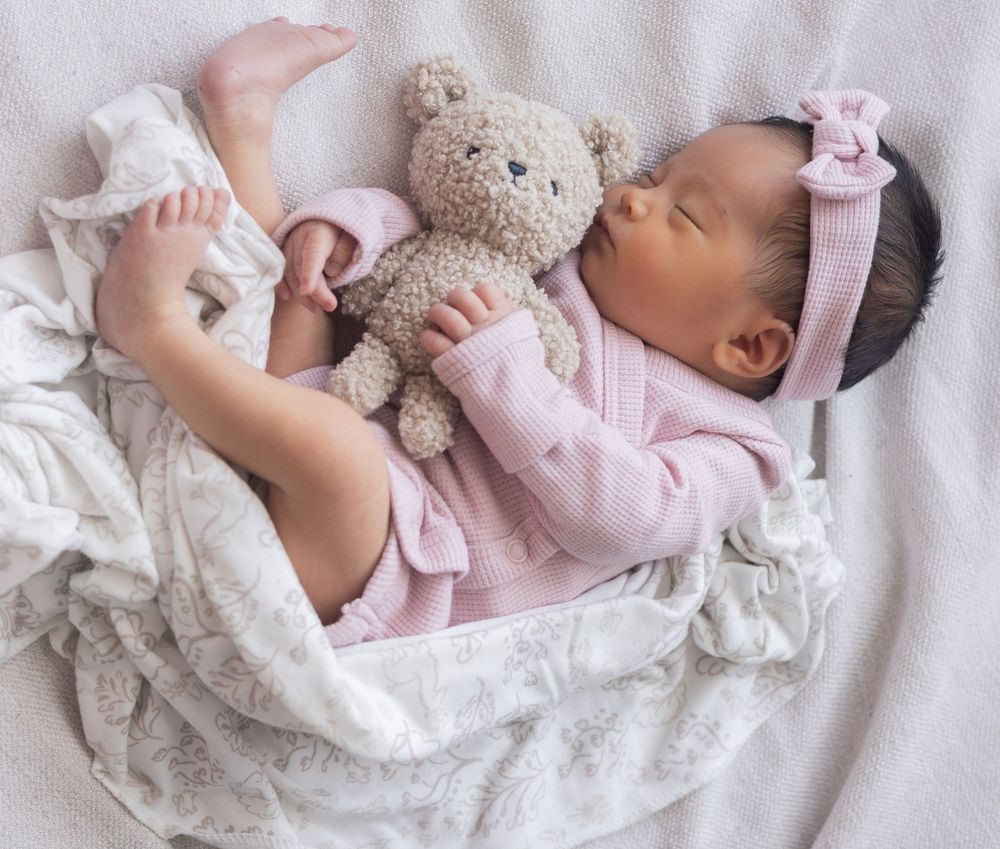 A sleeping baby in a soft pink outfit lies peacefully surrounded by cozy white and gray blankets.