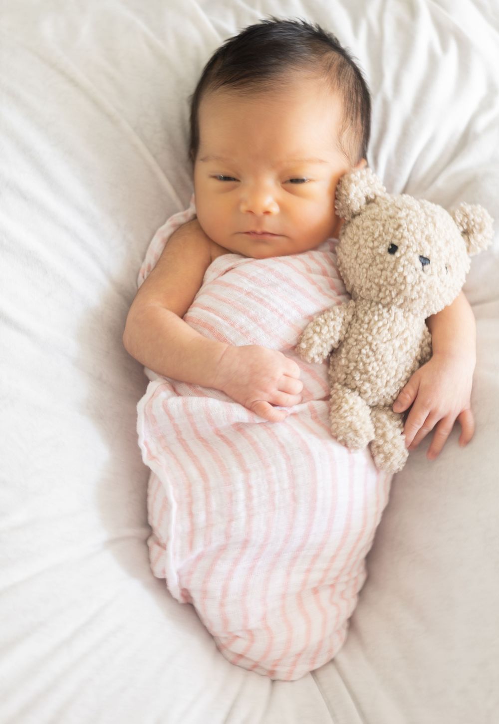 Sleeping infant in pink striped swaddle cuddles with plush teddy bear on white bedding.
