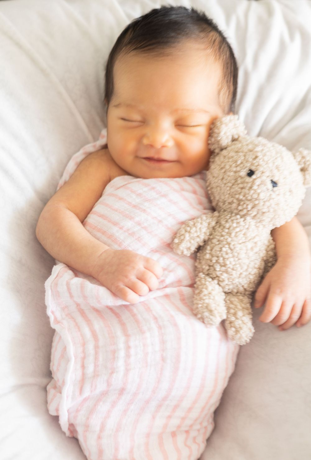 Peaceful sleeping baby in pink striped swaddle cuddling with soft beige teddy bear on white bedding.