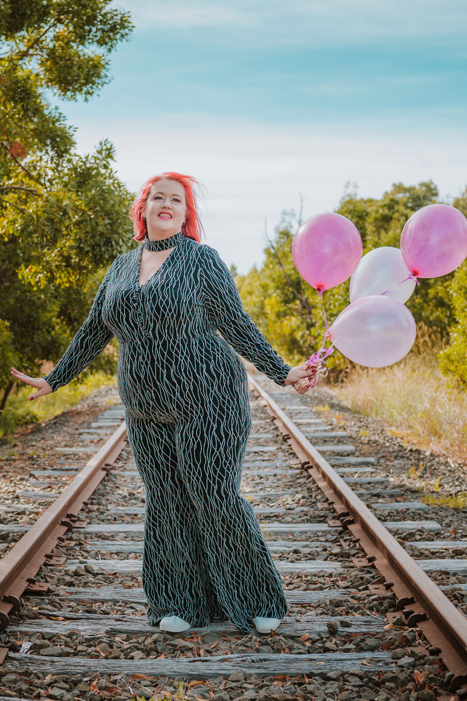 A figure in a dark dress stands on train tracks holding pink balloons against a blue sky on a sunny day.