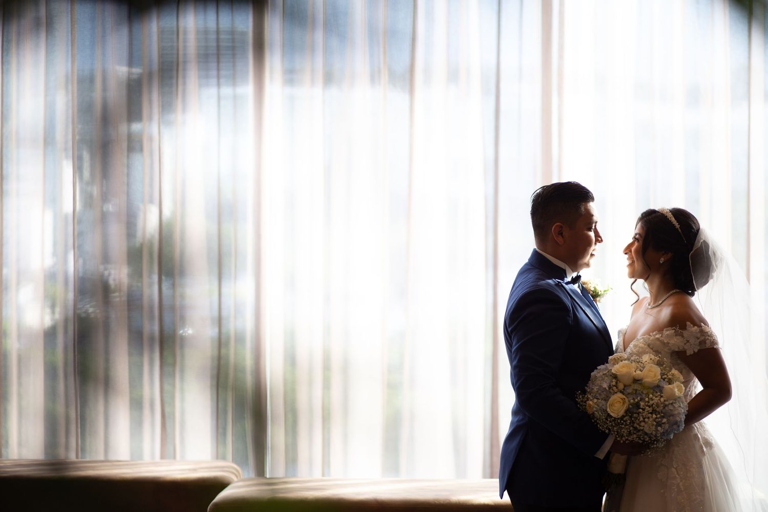 A romantic wedding couple silhouette against soft window light in an intimate indoor setting.