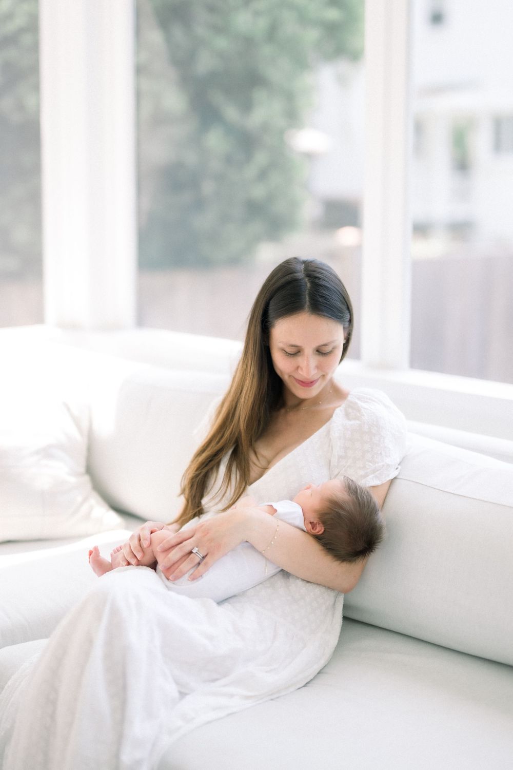 A tender moment captured on a white couch as a parent cradles a sleeping newborn baby in soft natural light.
