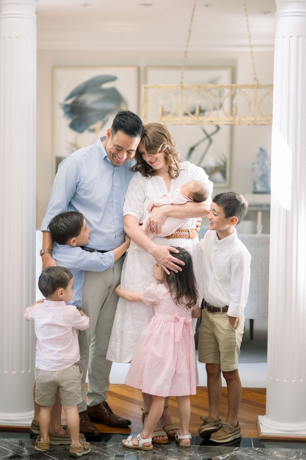 A tender family moment captured indoors with parents and their four children wearing light colored casual clothing.
