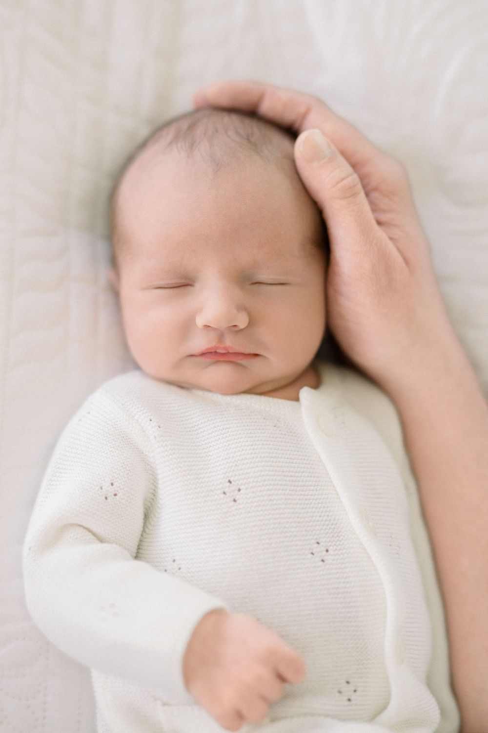 A sleeping newborn baby in white clothing is gently cradled by an adult's hand resting on their head.