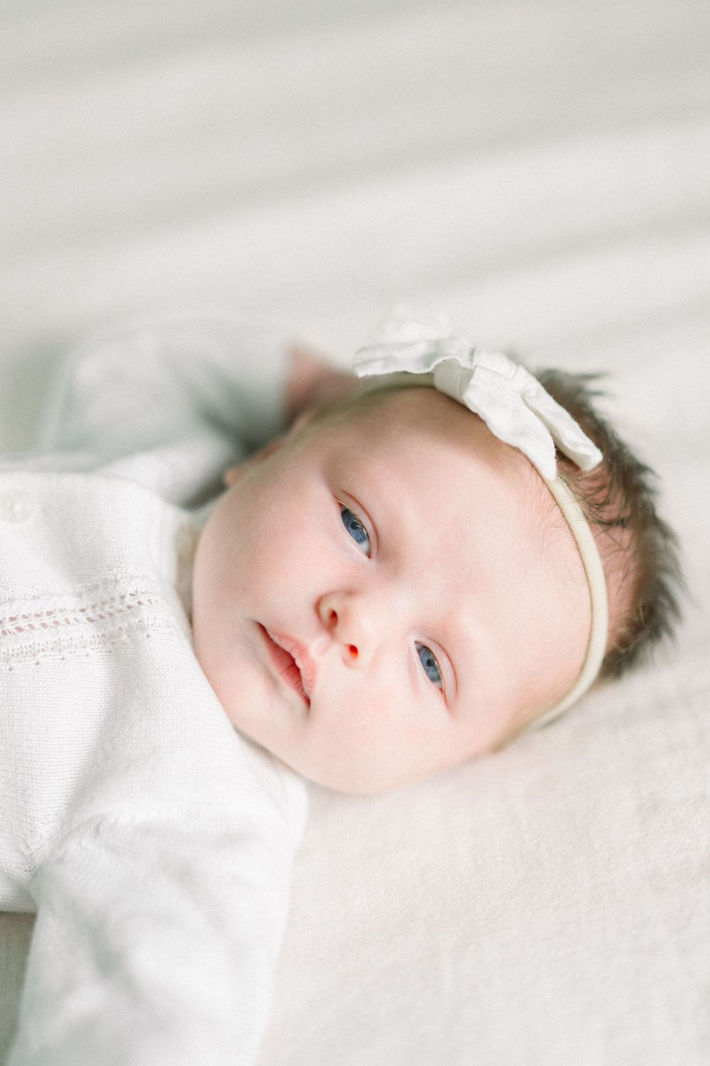 Sweet baby wearing white clothing and delicate bow headband rests peacefully on soft white bedding.