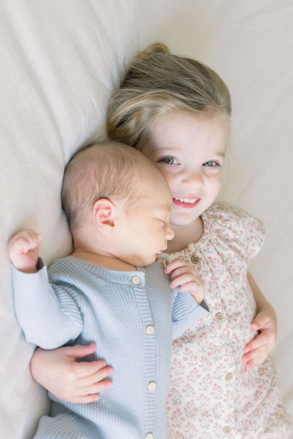 Two siblings share a tender moment lying together, with baby in blue cardigan and older child in floral dress.