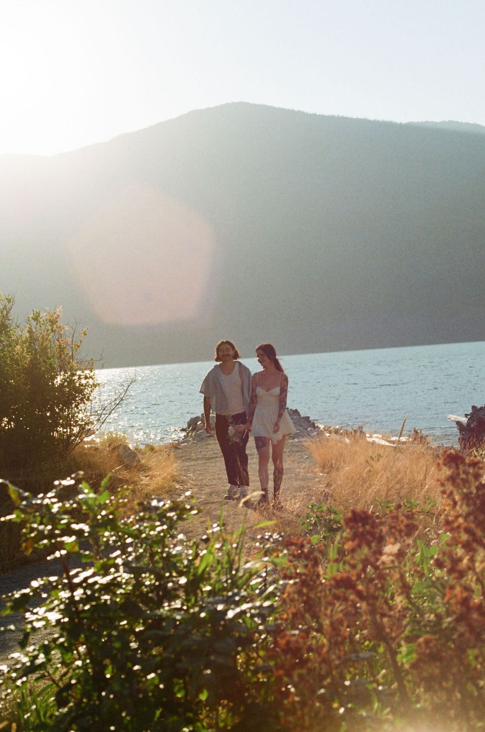 Couple walking along scenic coastal trail with wildflowers and mountains in golden sunset light.
