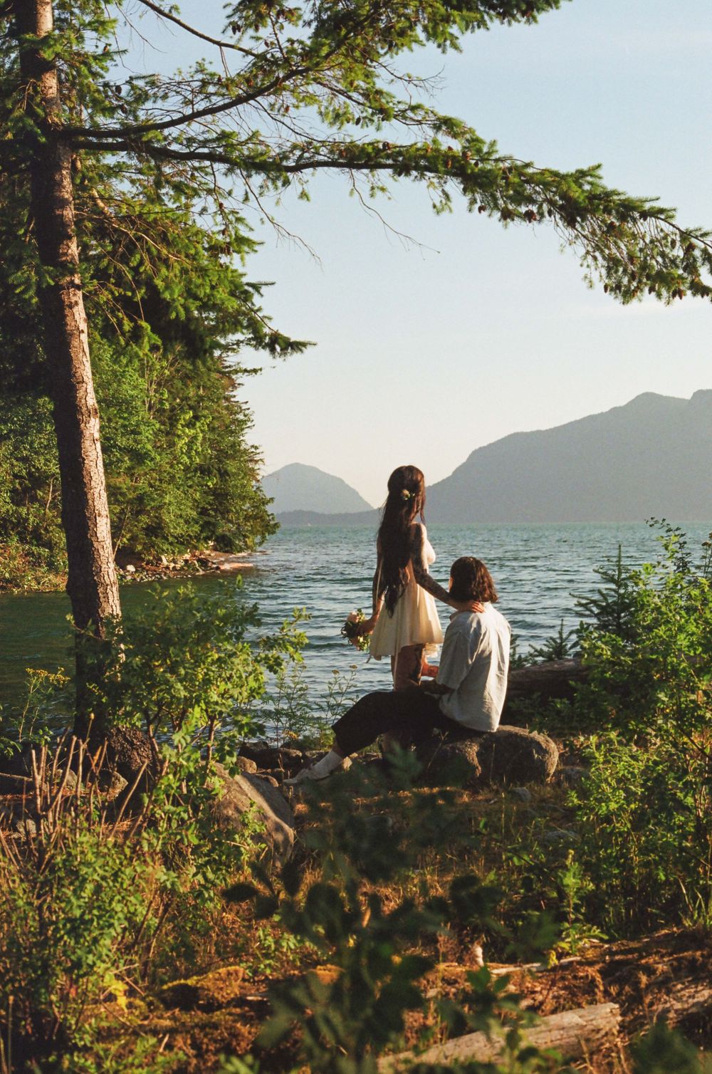 Two people sitting on rocky shore overlooking mountain lake at sunset.