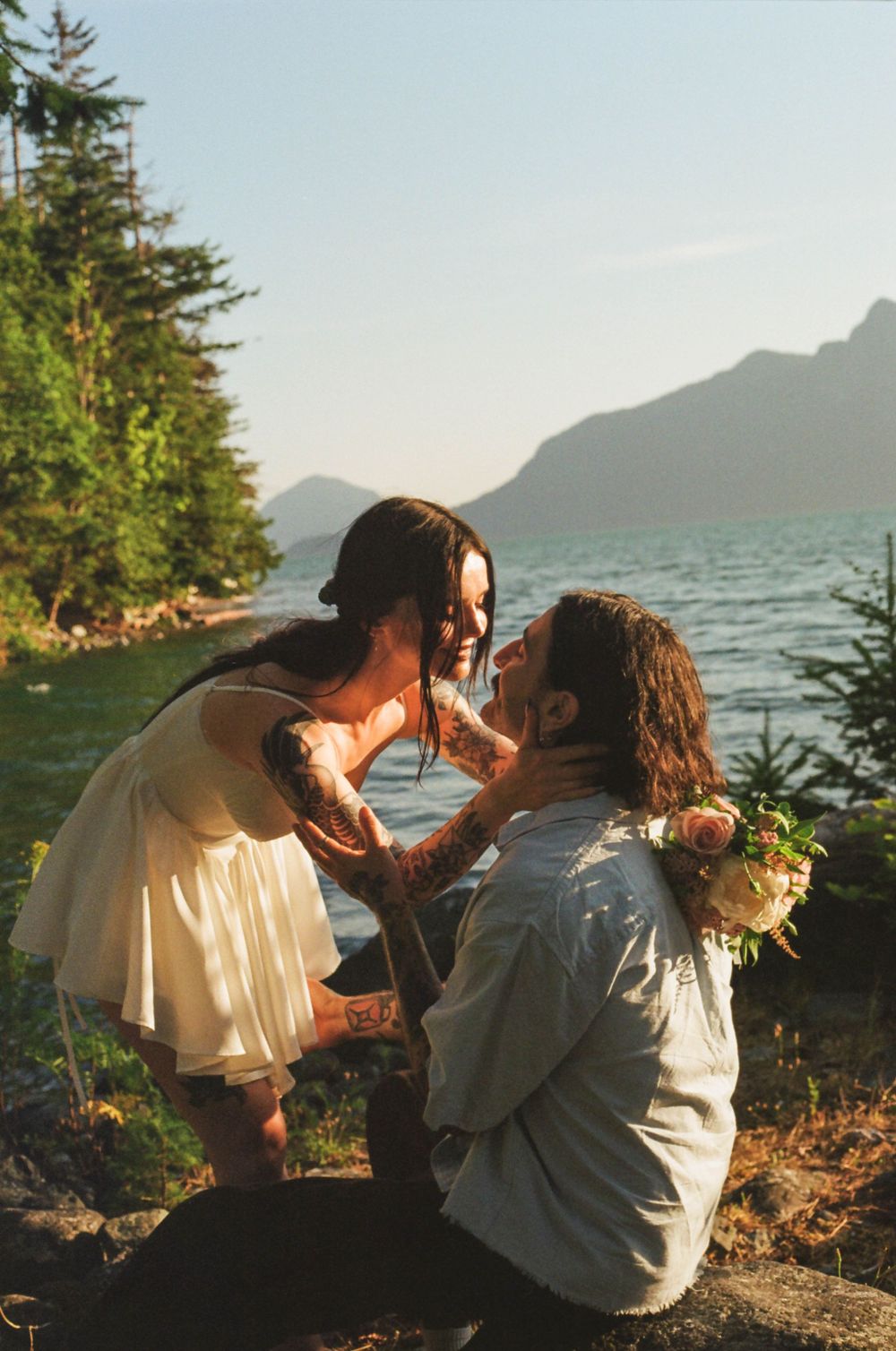 Couple sharing intimate moment on rocky shoreline with mountain lake backdrop.